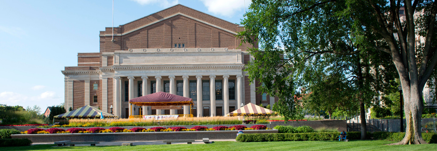 This photo shows Northrop Memorial Hall at the University of Minnesota. There is a green lawn, a large oak tree, and flowers in the signature U of M maroon and gold colors.