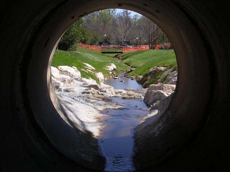 A shallow stream with rocks and grass on either side, as shot fromt he inside of a drain pipe