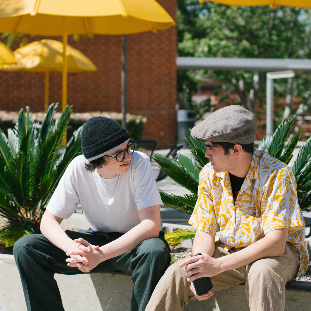 Two masculine presenting individuals sitting on a bench and talking.