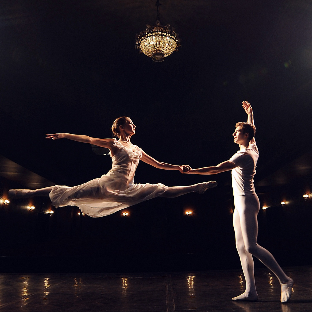 Two ballet dancers on stage, one standing and one mid-leap.