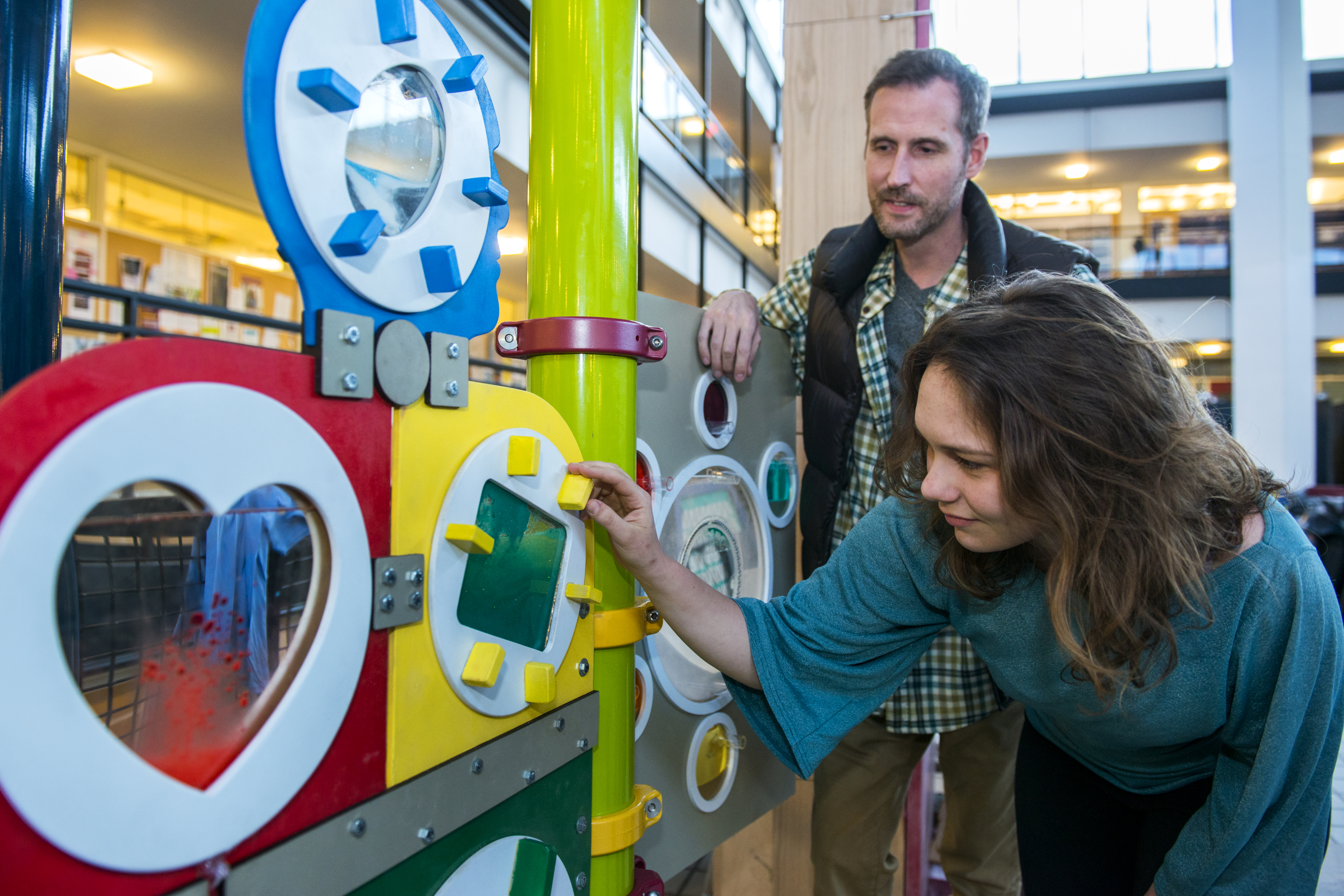 A woman playing with brightly colored gears on a wall while a man observes.