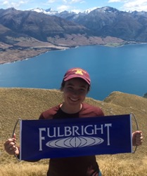 Photo of young woman holding Fulbright sign, standing in front of lake and mountains